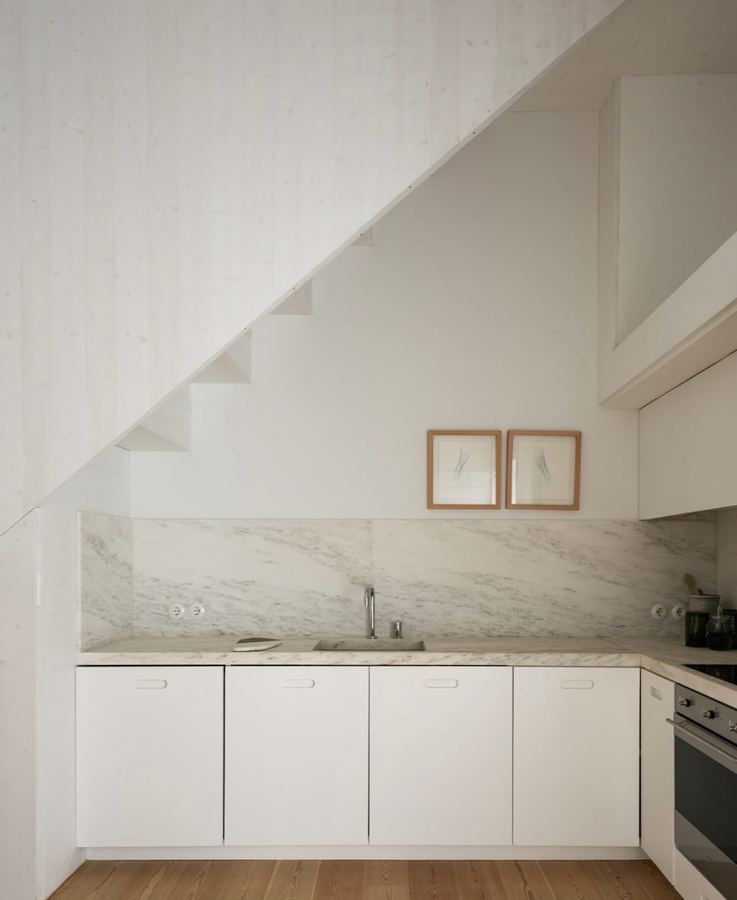 Compact white kitchen with Carrara marble backsplash floor-to-ceiling, white handleless cabinets with cut-out finger pulls, light wood floor, two small framed artworks