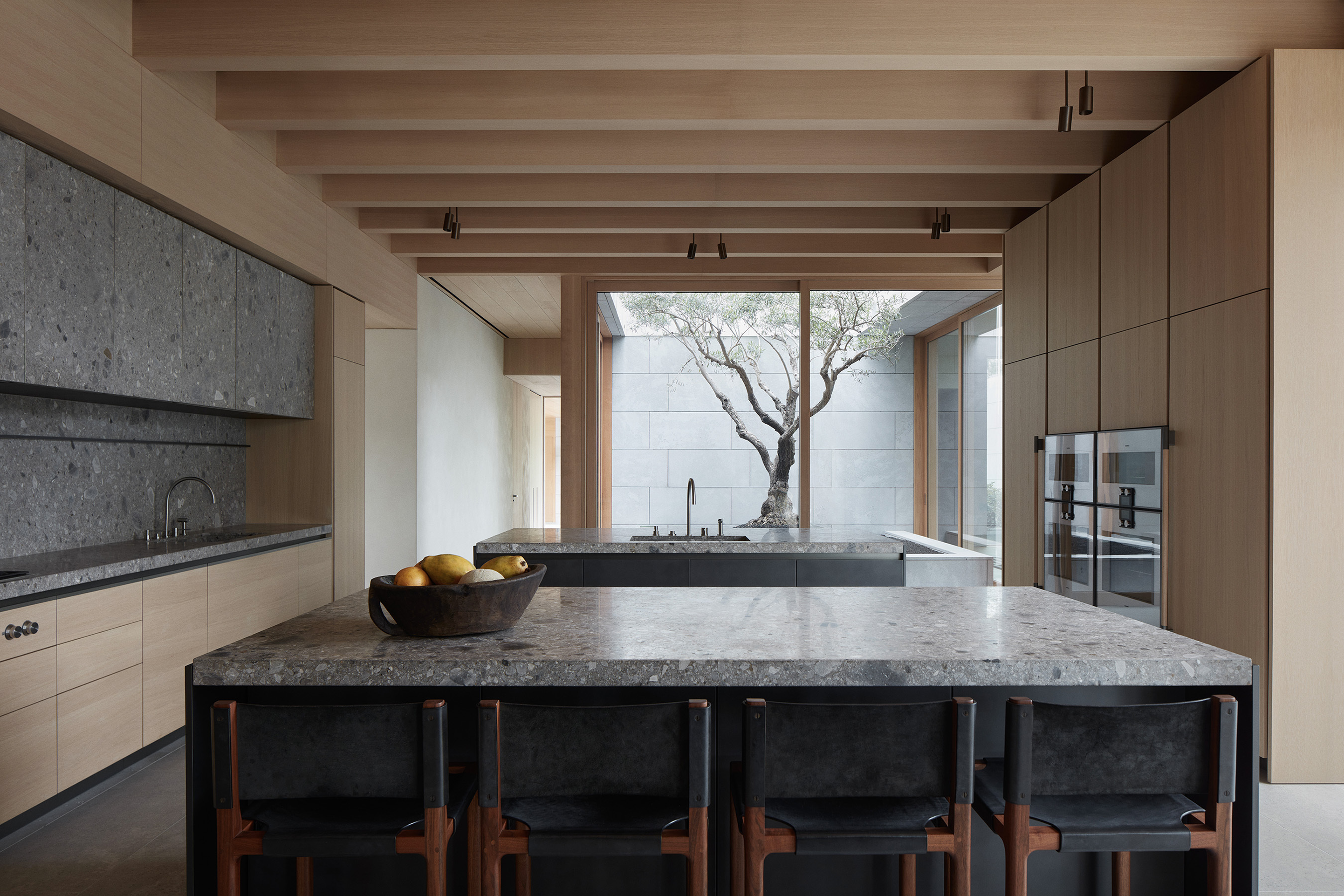 Vincent Van Duysen AN Residence kitchen interior: warm light-oak handleless cabinetry along both side walls and exposed oak ceiling beams above, central island with a dark grey terrazzo stone top and dark base, four leather-and-wood backed stools at the island, integrated wall ovens, central skylight opening over a mature olive tree against a pale stone courtyard wall