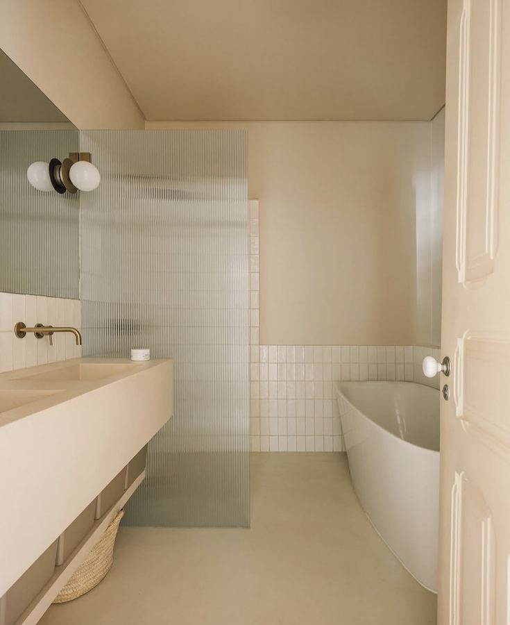 Cream microcement spa bathroom, monolithic poured vanity with double sink, brass faucets, reeded glass shower partition, small white tile shower wainscot, freestanding curved tub, mixed sconces in black and brass
