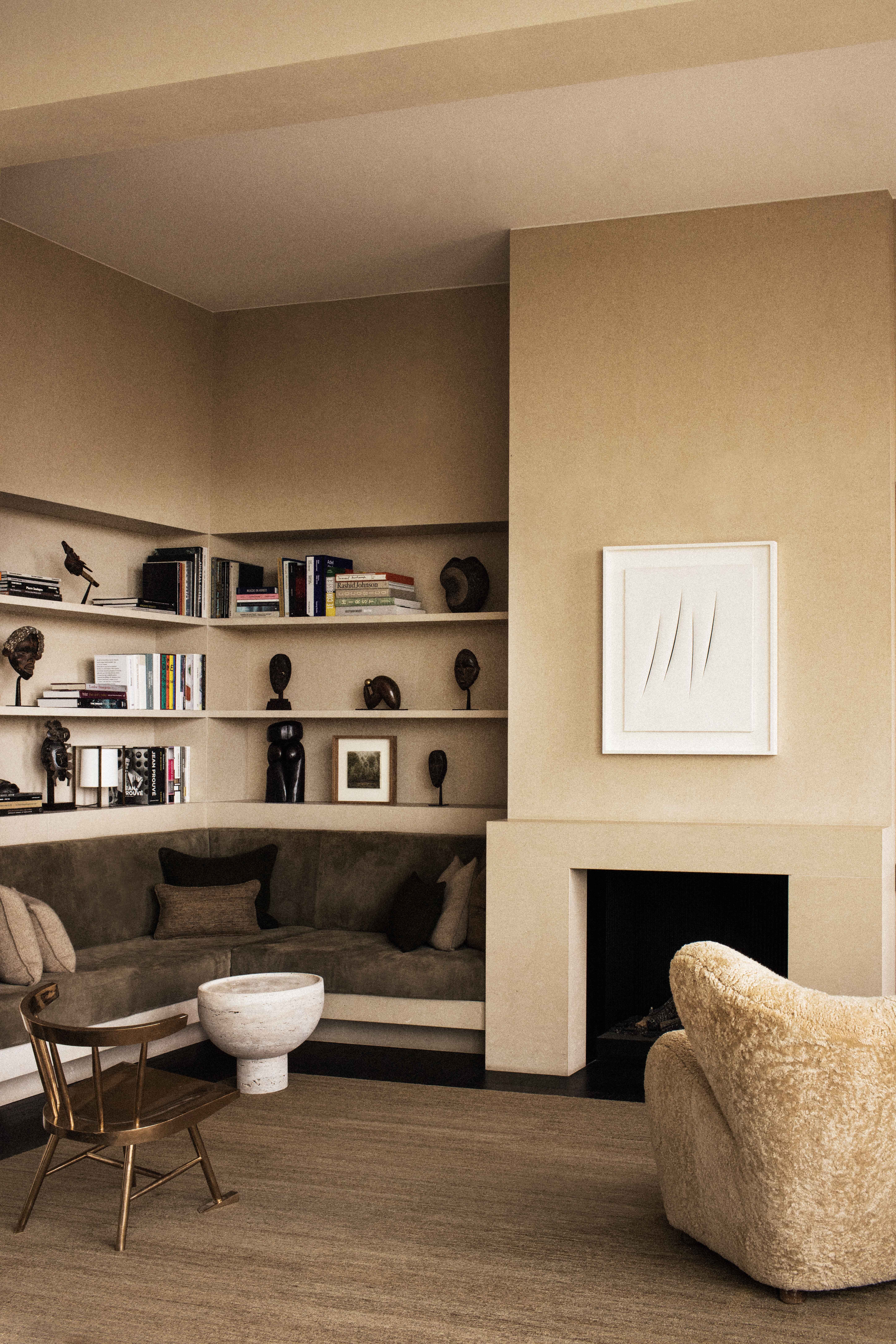 VVD RE Residence library corner: warm ochre-cream plaster walls, sculpted cream plaster fireplace surround with a small Fontana-style framed canvas above, built-in cream plaster banquette upholstered in dark olive-brown suede, continuous cream plaster floating shelves above holding African sculpture, books and small art, a cream sheepskin / bouclé armchair, a small brass tripod chair, a travertine footed bowl as side table, dark espresso oak floor, sisal area rug