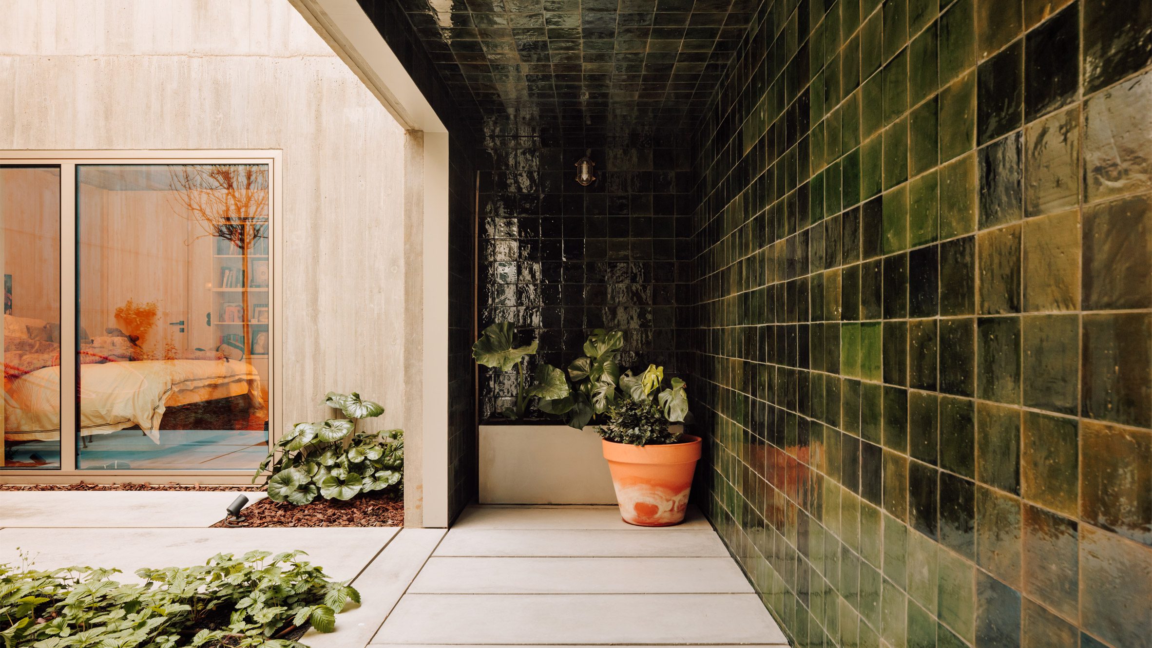 Bak Gordon Lisbon house courtyard: raw concrete wall on the left, dark glossy green-glazed tiles wrapping the right wall and the ceiling, large-format light grey concrete pavers as floor, terracotta planter with an elephant-ear plant, sliding glass door to a bedroom on the left, ground-cover planting outside